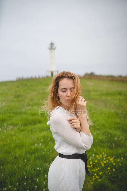 Mulher gentil gengibre em vestido branco com olhos fechados posando no prado verde no fundo do farol em Astúrias, Espanha — Fotografia de Stock