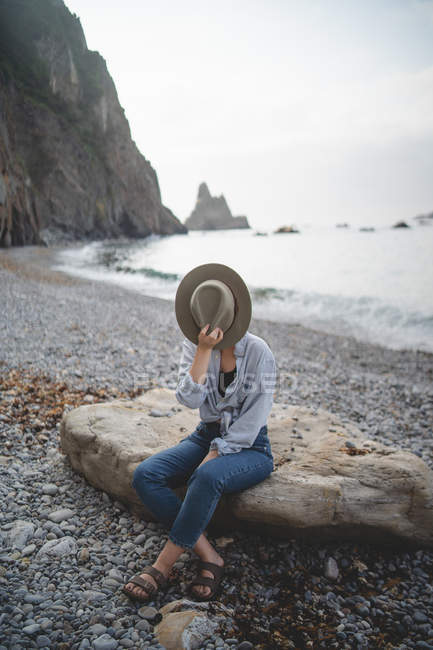 Tourist hiding face with hat enjoying seascape while chilling on big stone on rocky shore of Asturias looking away — Stock Photo