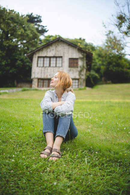 Mujer joven tranquila y casual disfrutando sentada en un prado verde cerca de una casa de madera en un pueblo rural en Asturias, España - foto de stock