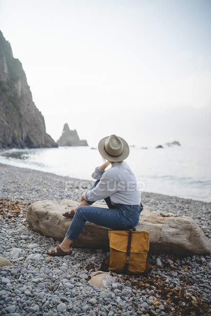 Жінка - туристка в капелюсі, яка насолоджується морською милицею, коли холоне на великому камені на скелястому березі Астурії. — стокове фото