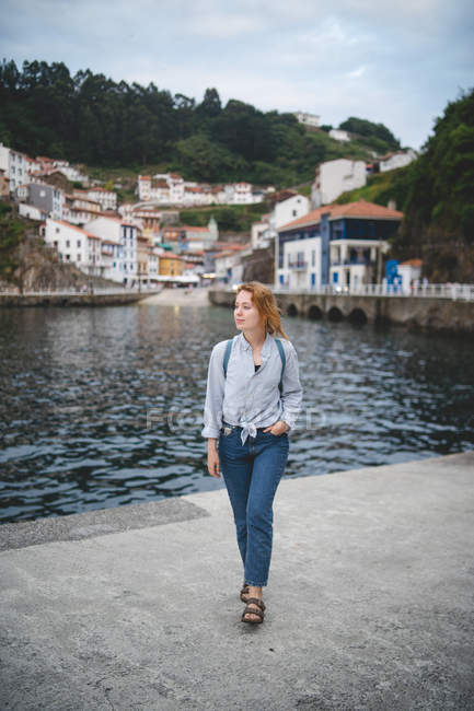 Donna con mano in tasca camminare su una banchina di cemento vicino all'acqua sullo sfondo della città nelle Asturie guardando altrove nelle Asturie, Spagna — Foto stock