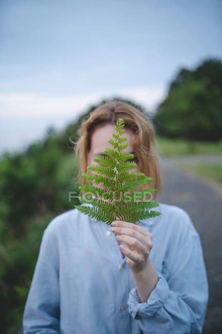 Rousse femme en chemise bleue couvrant le visage avec feuille de fougère verte et regardant à travers la nature des Asturies, Espagne — Photo de stock