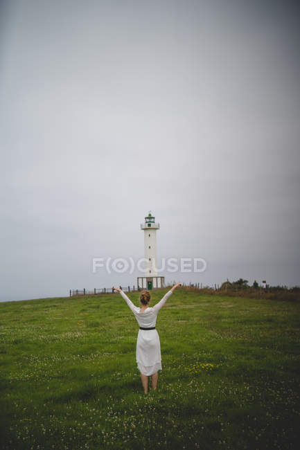 Rückansicht einer Frau im weißen Kleid, die bei bewölktem Wetter in Asturien, Spanien, nachdenklich im Feld gegen einen Leuchtturm geht — Stockfoto