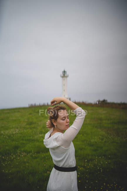 Rousse femme les yeux fermés en robe blanche marchant prudemment dans le champ contre le phare par temps nuageux dans les Asturies, Espagne — Photo de stock