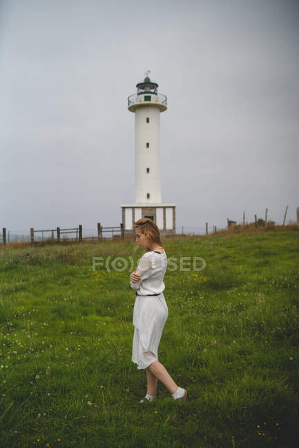 Vue latérale de la femme en robe blanche marchant prudemment dans le champ contre le phare par temps nuageux dans les Asturies, Espagne — Photo de stock