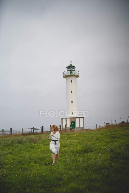 Femme rousse effrayée en robe blanche courant prudemment sur le terrain contre le phare par temps nuageux dans les Asturies, Espagne — Photo de stock