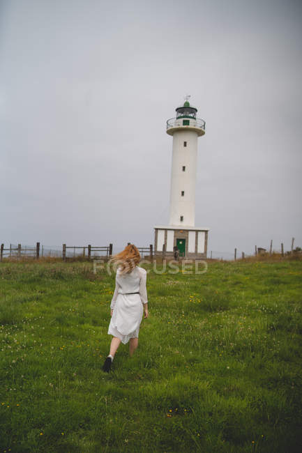 Rückansicht einer Frau im weißen Kleid, die bei bewölktem Wetter in Asturien, Spanien, nachdenklich im Feld gegen einen Leuchtturm geht — Stockfoto