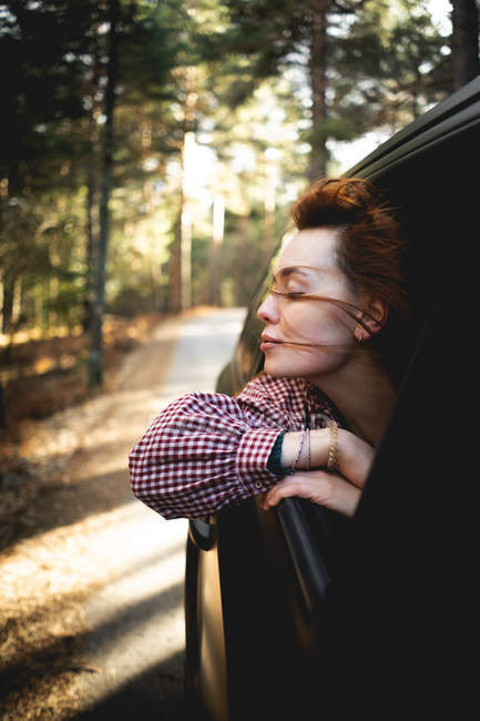 Vue latérale de la femme profitant du vent et de la liberté tout en sautant par la fenêtre de la voiture lors de l'équitation sur la route forestière dans les Asturies, Espagne — Photo de stock
