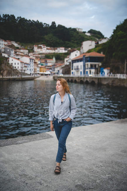 Frau mit der Hand in der Tasche, die am Betonkai in der Nähe des Wassers auf dem Hintergrund der Stadt in Asturien geht und in Asturien wegschaut, Spanien — Stockfoto