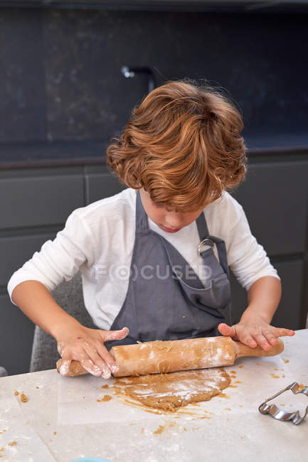 Кудрявый маленький ребенок в фартуке, стоящий и катящийся коричневое тесто с булавкой на столе на современной кухне — стоковое фото