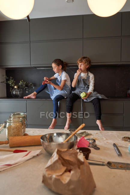 Niños satisfechos en delantales sentados en un armario de cocina moderno y comiendo frutas, mesa con herramientas de cocina en primer plano - foto de stock