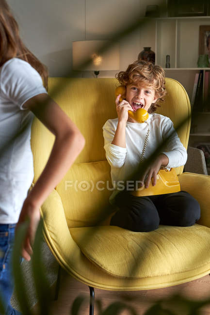 Niño con ropa casual sentado en un cómodo sillón amarillo y hablando por teléfono retro, chica al lado de salir de la habitación - foto de stock