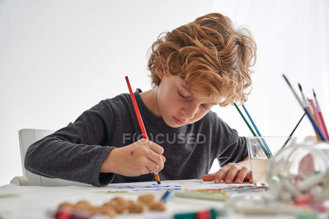 Petit garçon aux cheveux bouclés souriant assis à table et peignant contre un mur blanc à la maison — Photo de stock
