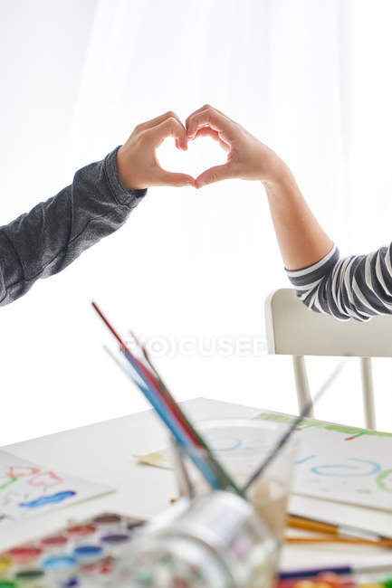 Маленька дівчинка і хлопчик посміхаються і показують жест серця, сидячи за столом і малюючи вдома разом. — стокове фото