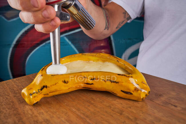 Sin rostro desde arriba hombre usando crema batidor y tuberías blanco crema fresca en plátano en la mesa - foto de stock