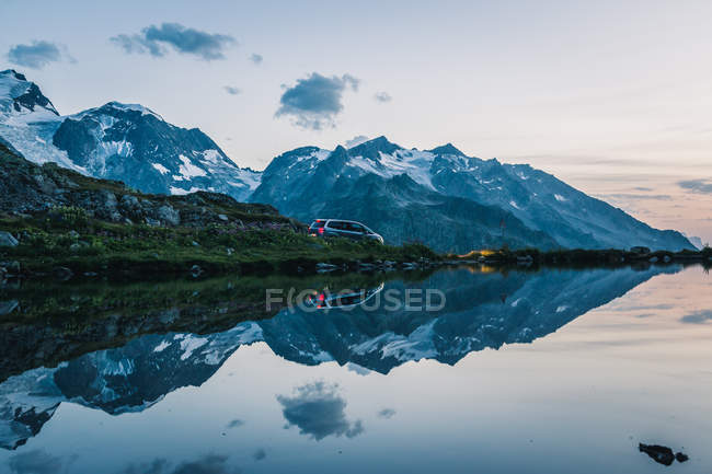 Voiture avec feux arrière sur la côte vide du lac calme dans les montagnes enneigées reflétant le ciel en Suisse — Photo de stock