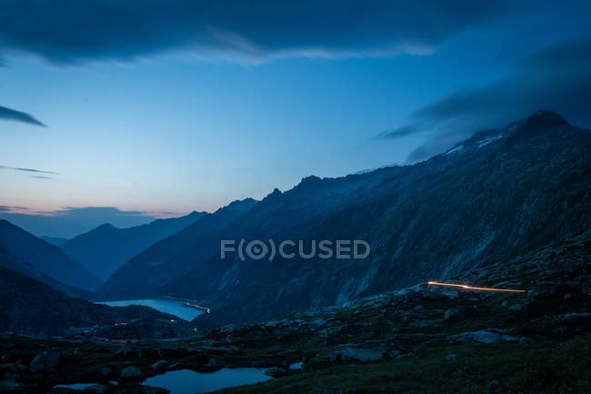 Таємничий темно-синій гірський хребет і річка між схилами з вогнями вздовж дороги в Швейцарії. — стокове фото