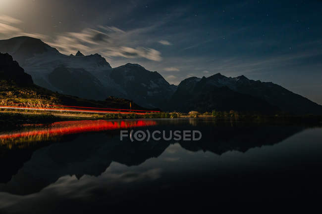 Impresionante paisaje dramático de orilla roja y aguas oscuras y tranquilas que reflejan el cielo nublado y las montañas en Suiza - foto de stock