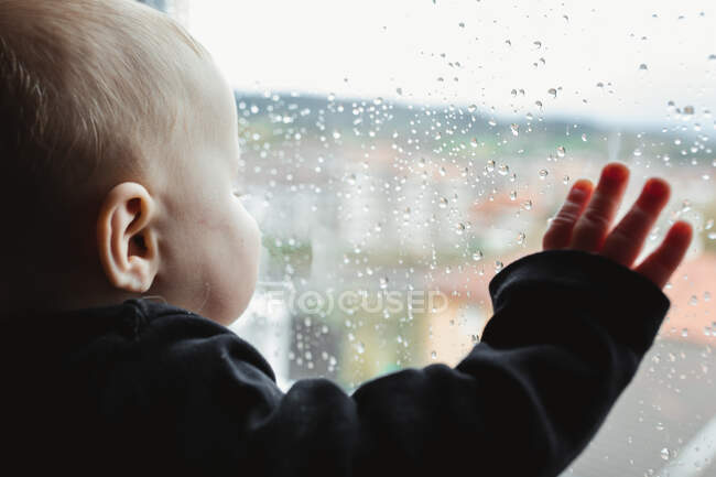 Bebé anónimo mirando por la ventana húmeda - foto de stock
