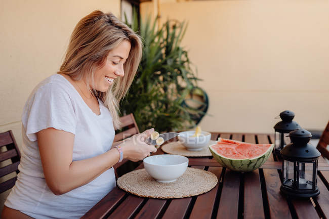 Vista lateral de la mujer embarazada en ropa de casa blanca cortando plátano a tazón blanco en la terraza - foto de stock