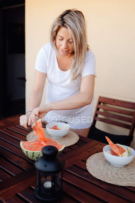 Pregnant woman in white home clothes cutting watermelon to white bowl at terrace — Stock Photo