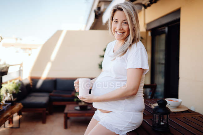 Cheerful blonde pregnant woman leaning on wooden table at terrace while drinking morning tea and looking at camera — Stock Photo
