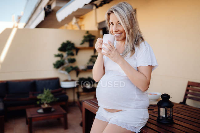 Alegre mujer embarazada rubia apoyada en la mesa de madera en la terraza mientras bebe té de la mañana y mira a la cámara - foto de stock