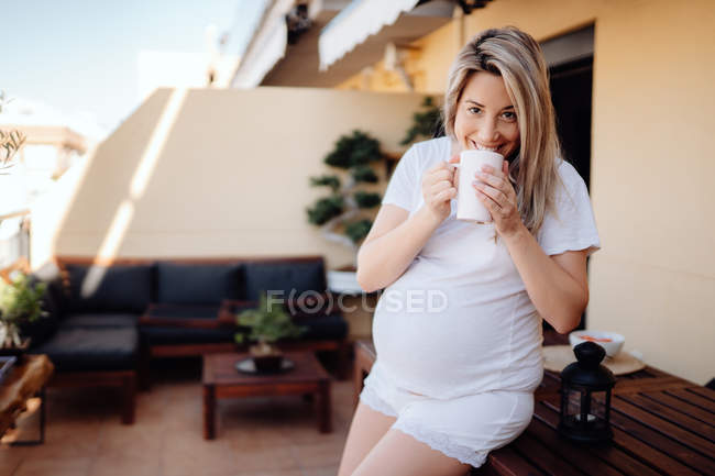 Allegro donna bionda incinta appoggiata sul tavolo di legno in terrazza mentre beve tè del mattino e guardando la fotocamera — Foto stock