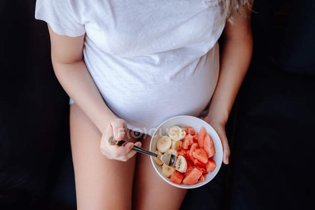 Imagem cortada da mulher grávida comendo bananas fatiadas e melancia da tigela com garfo enquanto se senta no sofá escuro — Fotografia de Stock