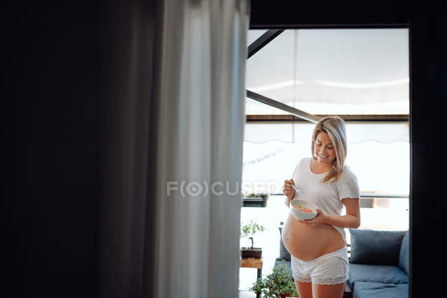 Content femme enceinte blonde calme debout à la maison contre les grandes fenêtres tenant bol à la main — Photo de stock