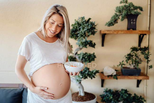 Вміст спокійна блондинка вагітна жінка стоїть вдома проти стіни з рослинами і торкається голого живота, тримаючи миску в руці — стокове фото