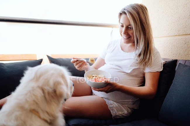 Cheerful blonde pregnant woman in white homey clothes feeding labrador dog with piece of banana from bowl while sitting on sofa at terrace — Stock Photo