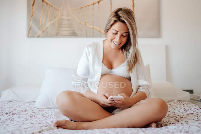 Содержание блондинка беременная женщина в белой открытой рубашке и лифчик просматривает мобильный телефон, сидя со скрещенными ногами на большой кровати в светлой комнате — стоковое фото