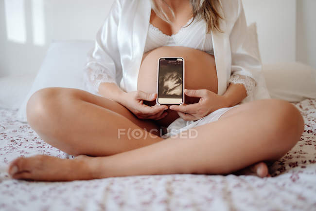 Cropped image of pregnant woman demonstrating picture of ultrasound scan on smartphone while sitting in bra and open shirt — Stock Photo