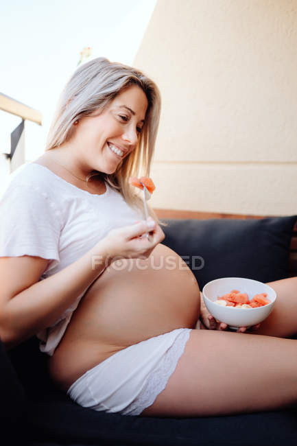 Pregnant woman wearing white t shirt eating sliced bananas and watermelon from bowl with fork while sitting on dark sofa — Stock Photo