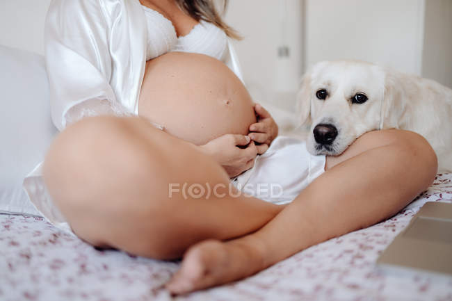 Geschnittenes Bild einer schwangeren Frau, die mit gekreuzten Beinen auf dem Bett sitzt und den Bauch berührt — Stockfoto