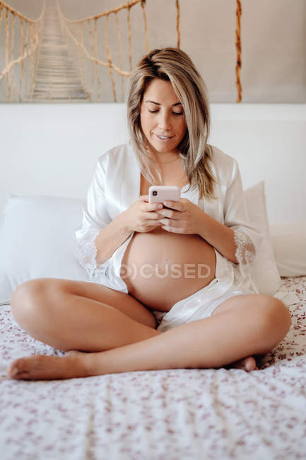 Содержание блондинка беременная женщина в белой открытой рубашке и лифчик просматривает мобильный телефон, сидя со скрещенными ногами на большой кровати в светлой комнате — стоковое фото