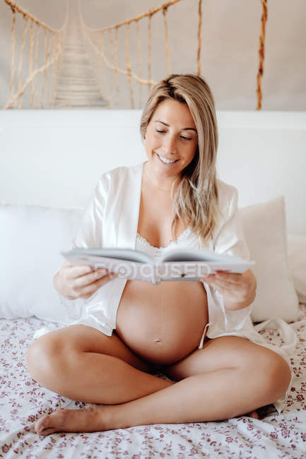 Zufriedene blonde schwangere Frau in Hauskleidung liest Buch, während sie mit überkreuzten Beinen auf dem Bett im hellen Zimmer sitzt — Stockfoto