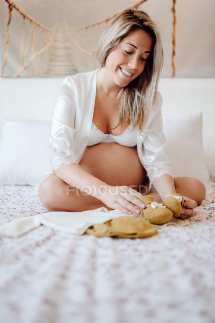 Mujer embarazada vestida con blusa blanca abierta y sujetador mostrando ropa para el feto mientras está sentada en la cama con las piernas cruzadas - foto de stock