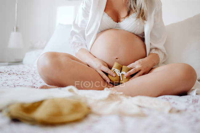 Cropped image of pregnant woman dressed in white open blouse and bra showing clothes for unborn while sitting on bed with crossed legs — Stock Photo