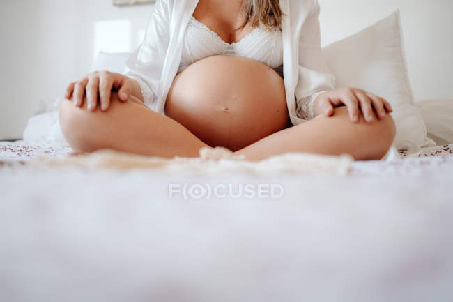 Image recadrée d'une femme enceinte vêtue d'une blouse blanche ouverte et d'un soutien-gorge assis sur un lit avec des jambes croisées — Photo de stock