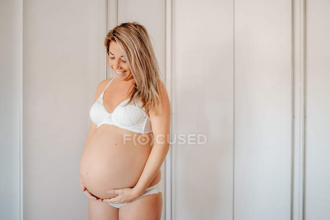 Happy blonde pregnant woman dressed in white bra and panties holding belly while standing against bright wall — Stock Photo
