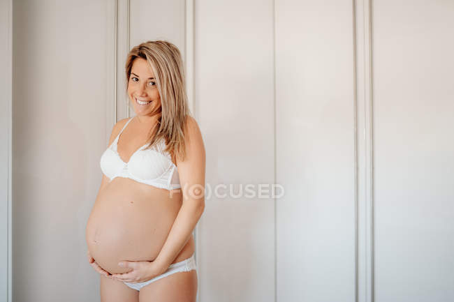 Glückliche blonde schwangere Frau in weißem BH und Höschen, die den Bauch hält, während sie an der hellen Wand steht und in die Kamera schaut — Stockfoto