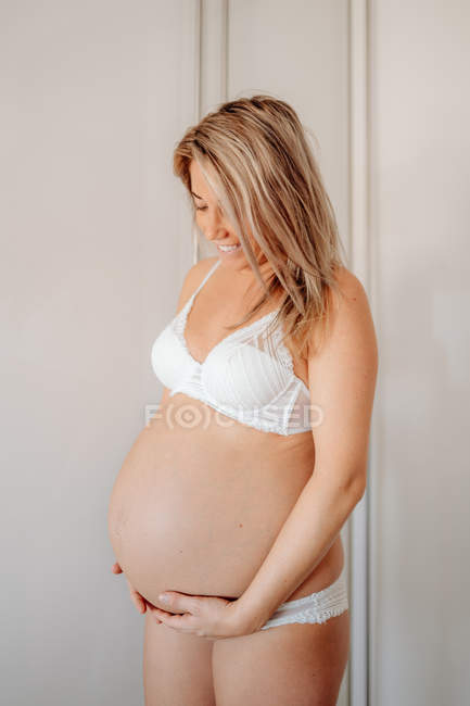 Mujer embarazada rubia feliz vestida con sujetador blanco y bragas sosteniendo el vientre mientras está de pie contra la pared brillante - foto de stock
