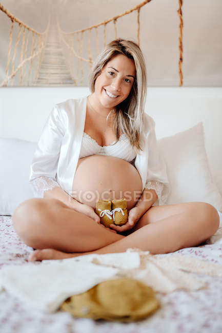 Femme enceinte vêtue d'une blouse blanche ouverte et d'un soutien-gorge montrant des vêtements pour enfants à naître assis sur un lit avec des jambes croisées — Photo de stock