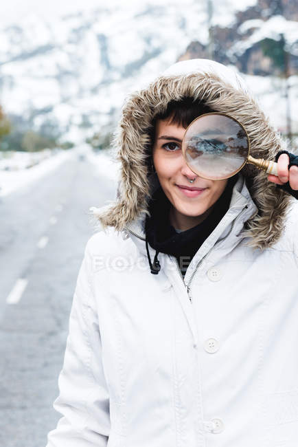 Donna con piercing al naso indossa giacca bianca invernale con cappuccio sulla testa guardando la fotocamera attraverso lente d'ingrandimento — Foto stock
