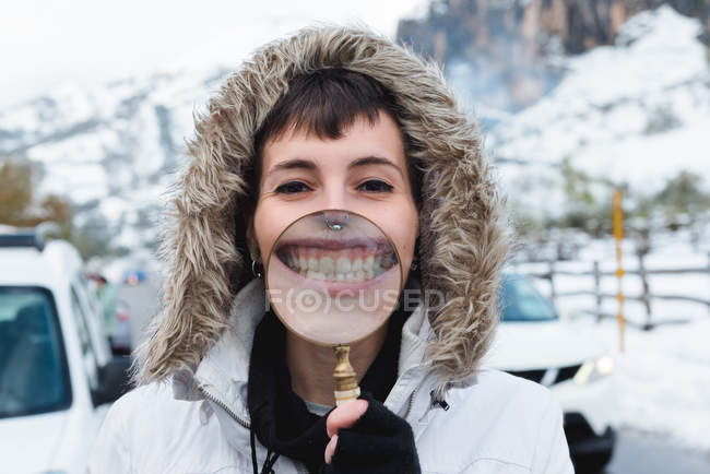 Woman with nose piercing in white winter jacket with hood on head looking at camera smiling teeth throughout magnifying glass — Stock Photo
