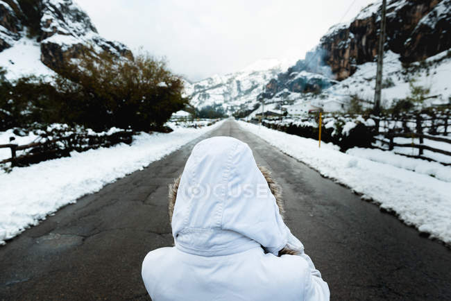 Rückansicht einer Person in weißer Winterjacke mit Kapuze auf dem Kopf, die mitten auf einer asphaltierten Straße am Fuße der Berge bei verschneitem Wetter steht — Stockfoto