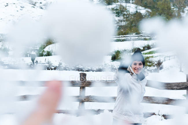 Женщина в черной вязаной шляпе с помпоном и белой зимней курткой бросает снежок в камеру во время прогулки в горной деревне — стоковое фото
