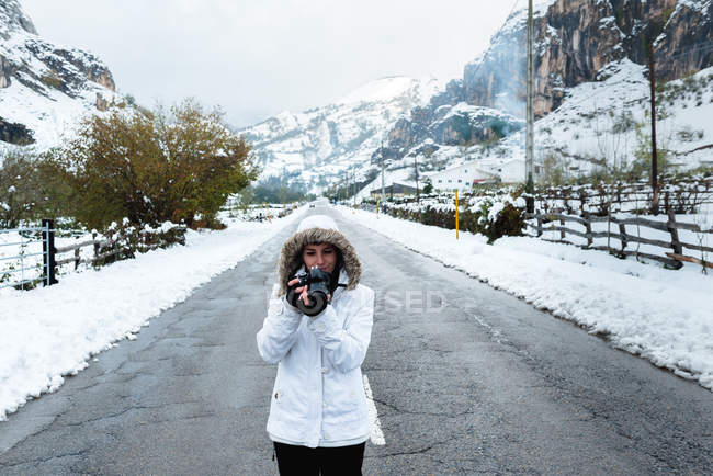 Frau in weißer Winterjacke mit Kapuze und schwarzer Hose fotografiert mit Kamera mitten auf asphaltierter Straße — Stockfoto